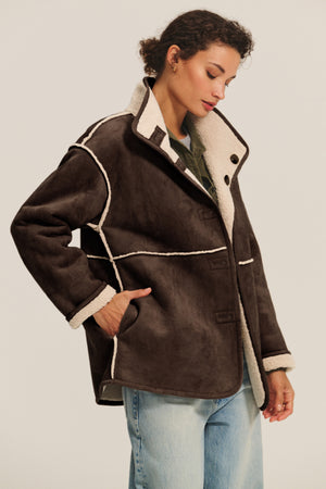 A woman wears the Velvet by Graham & Spencer SHONA SHERPA REVERSIBLE JACKET with cream trim over a green top and light blue jeans, standing against a plain background.