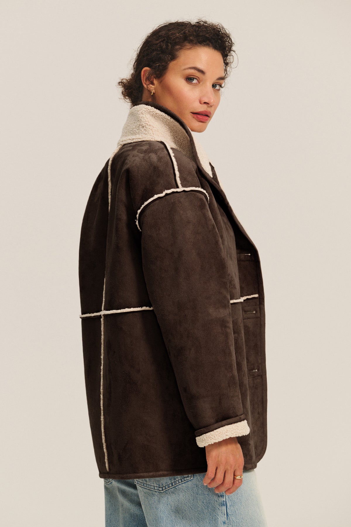   A woman with curly hair, shown in profile against a plain background, wears the SHONA SHERPA REVERSIBLE JACKET by Velvet by Graham & Spencer—a brown faux suede, shearling-lined style—paired with light blue jeans. 