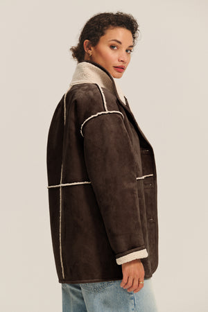 A woman with curly hair, shown in profile against a plain background, wears the SHONA SHERPA REVERSIBLE JACKET by Velvet by Graham & Spencer—a brown faux suede, shearling-lined style—paired with light blue jeans.