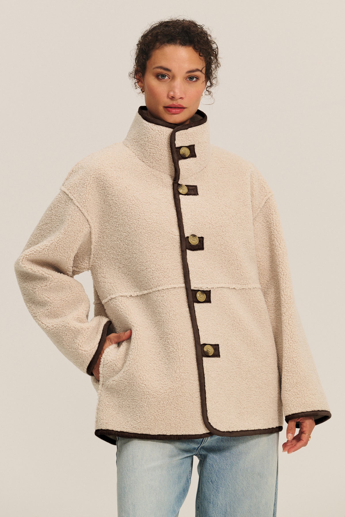 A woman wears the SHONA SHERPA REVERSIBLE JACKET by Velvet by Graham & Spencer, a cream, high-collar jacket with faux sherpa, dark trim, and large buttons, paired with light blue jeans against a plain background.-39814926008513