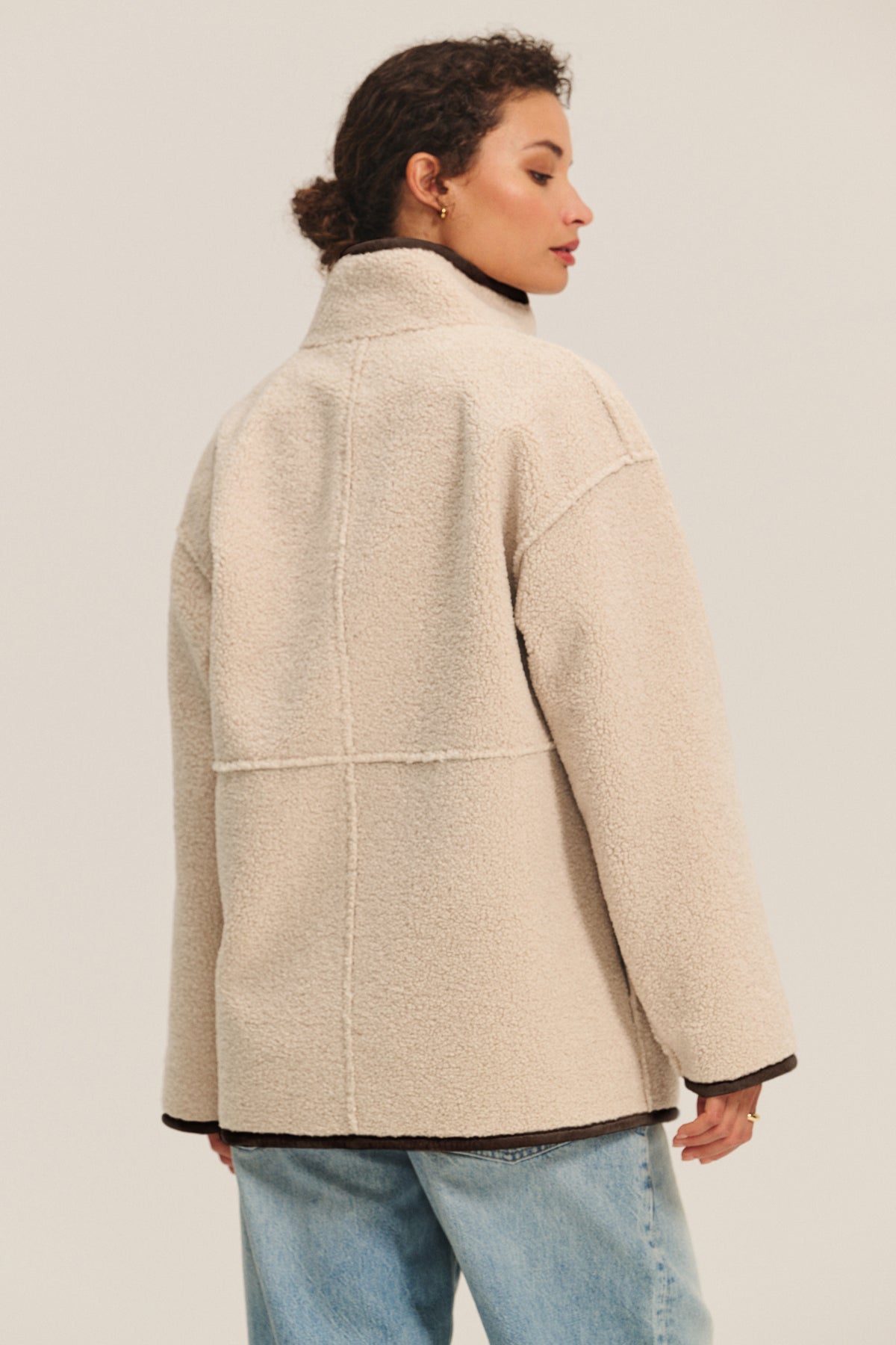   A person is seen from the back wearing the SHONA SHERPA REVERSIBLE JACKET by Velvet by Graham & Spencer in cream, paired with light blue jeans, against a plain background. 