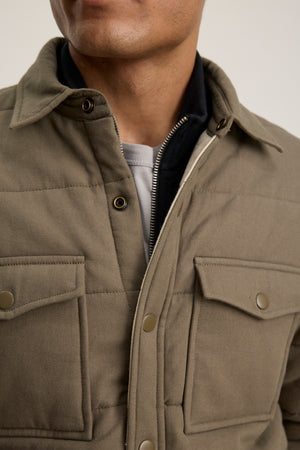 A person wearing the SUMMIT JACKET by Velvet by Graham & Spencer—a khaki quilted cotton jacket with a snap-button front, layered over a zip-up piece and a grey shirt underneath.