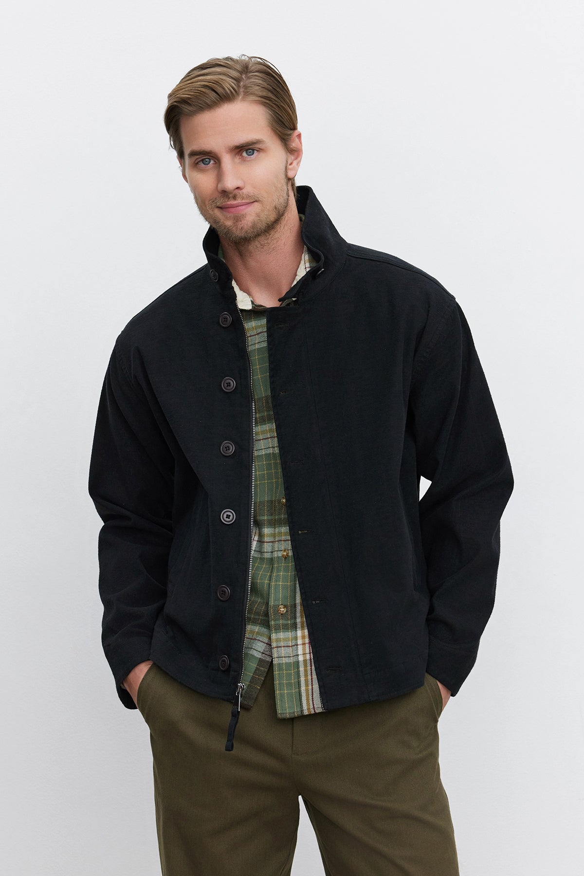   A man stands with hands in pockets, wearing the WILBUR JACKET by Velvet by Graham & Spencer over a plaid shirt and green pants. The black jacket, crafted from a brushed cotton blend twill, complements his neutral expression as he looks directly at the camera. 