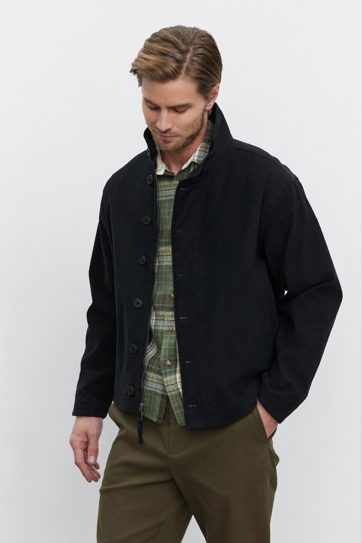   A man with light brown hair wearing a Velvet by Graham & Spencer WILBUR JACKET over a green plaid, button-front shirt and olive green twill pants stands against a plain white background. He is looking down with one hand in his pocket. 