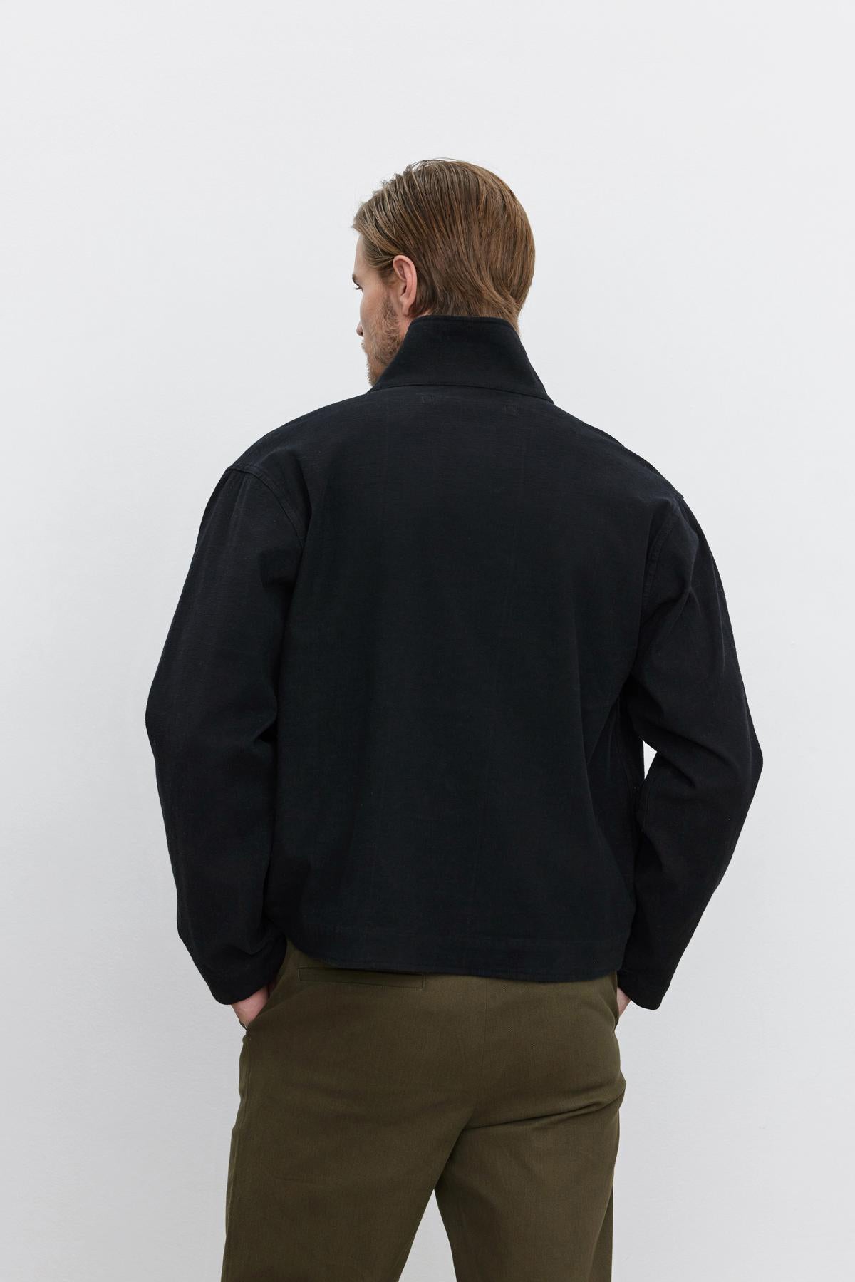   A person with light brown hair is seen from behind, wearing the WILBUR JACKET by Velvet by Graham & Spencer and olive green twill pants with a button front, standing against a plain white background. 