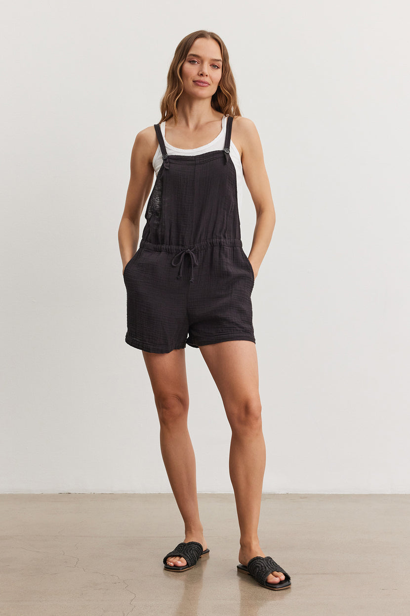 A woman stands in a studio, wearing a black CYNDEE COTTON GAUZE ROMPER by Velvet by Graham & Spencer with drawstring waist and black slide sandals, smiling subtly.