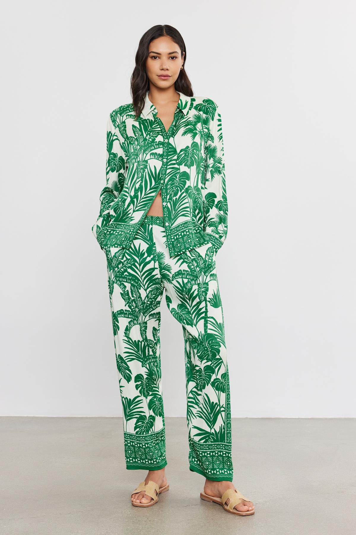  A woman wearing a green and white palm print pantsuit from Velvet by Graham & Spencer, with a matching crop top, standing against a plain backdrop, paired with beige sandals. 