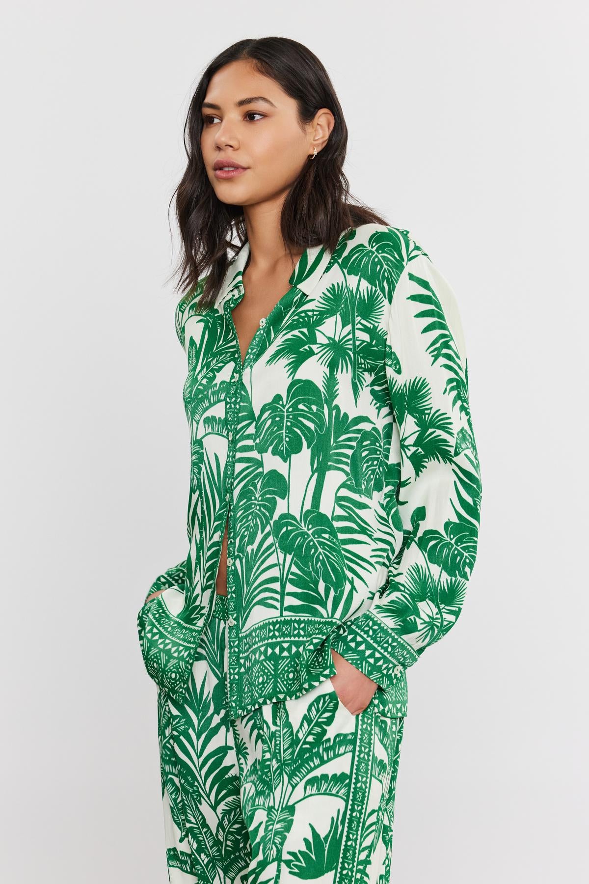 A woman stands wearing a green and white palm print AYLA BUTTON-UP SHIRT pajama set by Velvet by Graham & Spencer, looking to the side, with a calm expression.-36910265073857