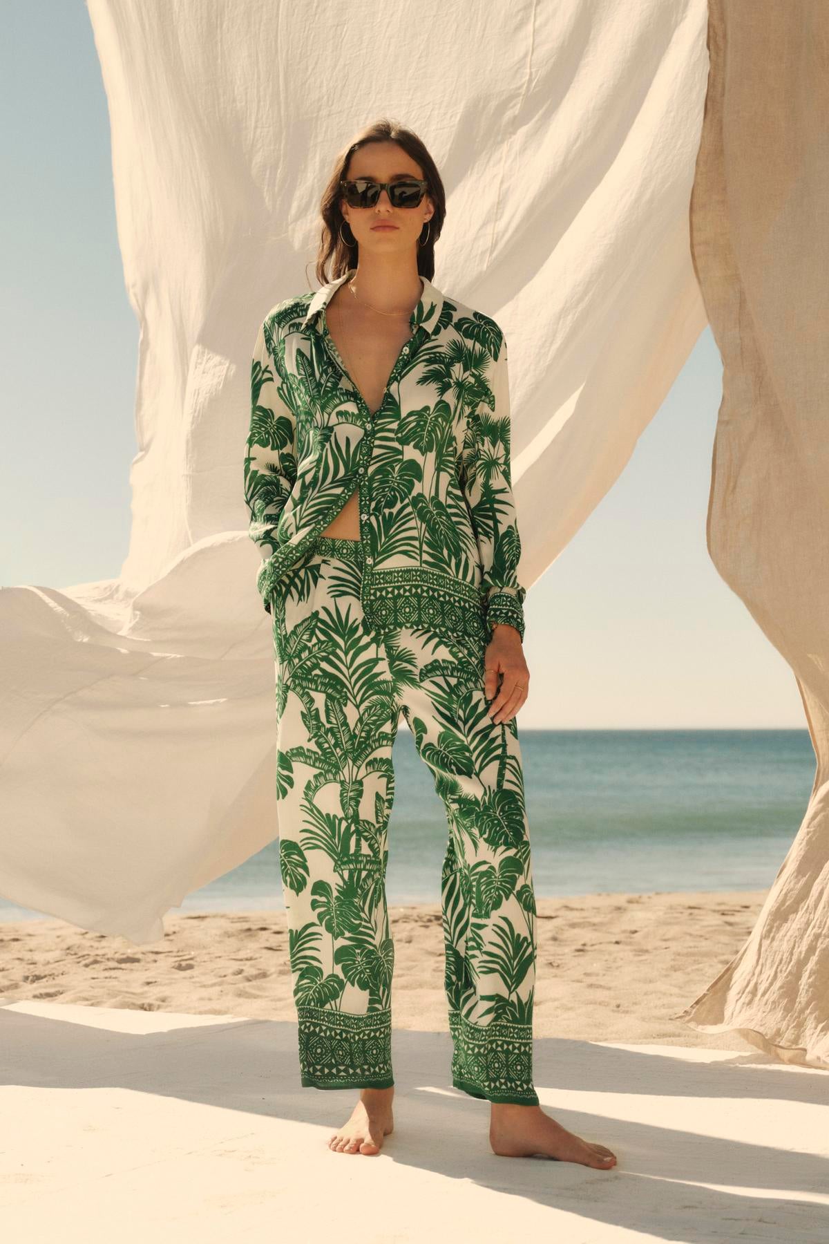   A woman in a green Velvet by Graham & Spencer AYLA button-up shirt stands on a beach, flanked by billowing white fabrics, with sunglasses on. 
