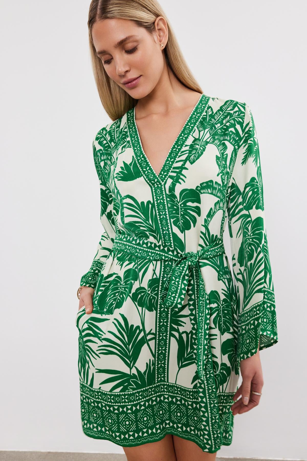Woman wearing a Velvet by Graham & Spencer EMELLA DRESS, a green and white palm print dress with a v-neckline and detachable belt, posing in a studio setting.-36917044052161