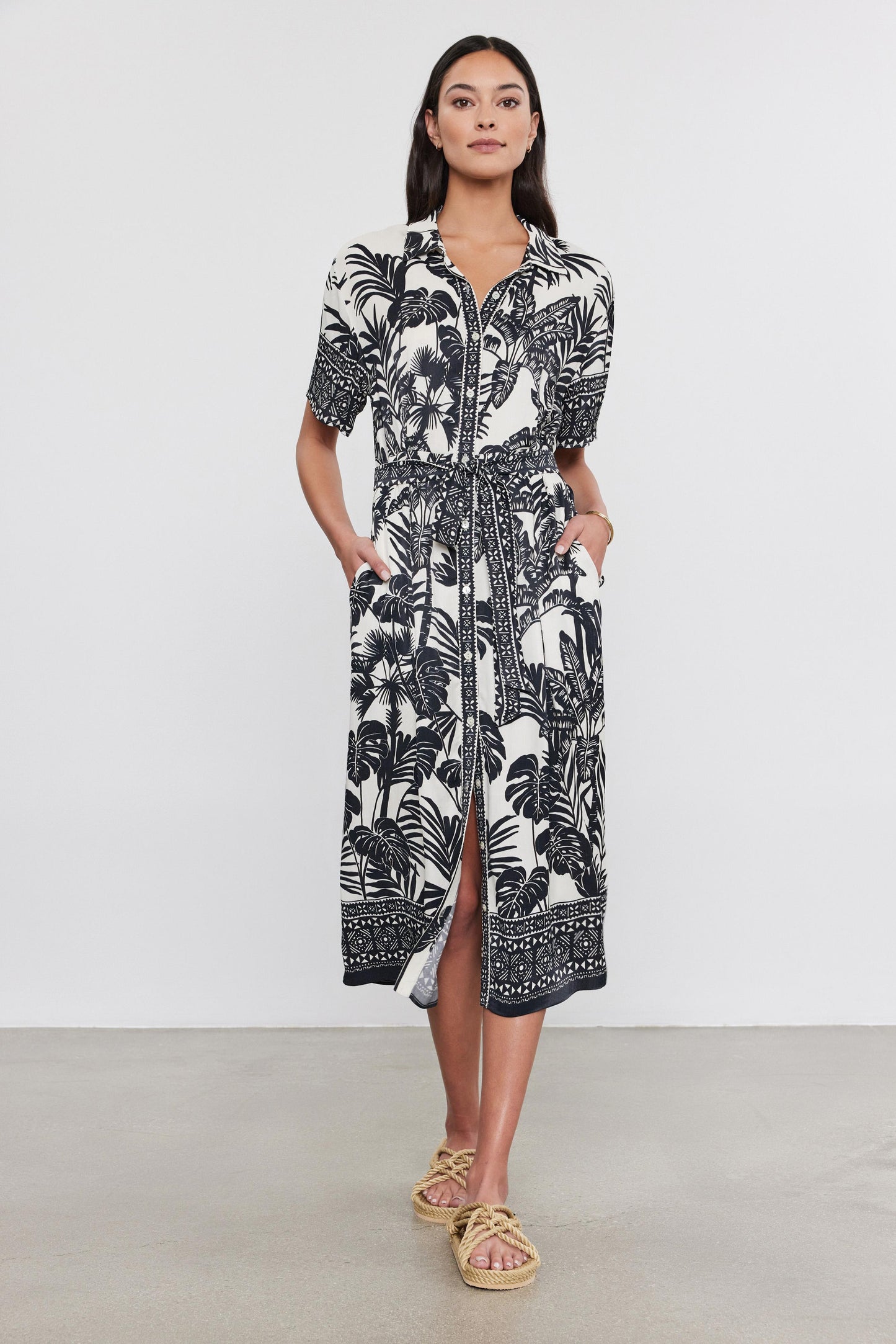 A woman stands in a studio wearing a black and white palm print Freya Dress by Velvet by Graham & Spencer with a front zipper and sandals.-36910269792449