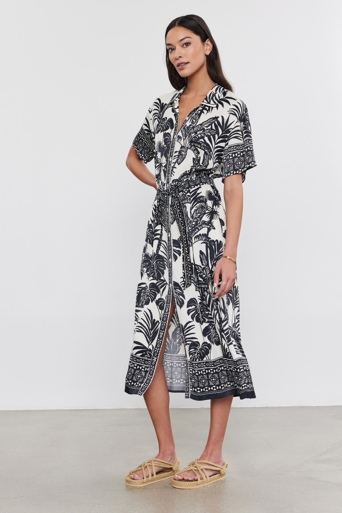A woman in a black and white palm print Velvet by Graham & Spencer FREYA DRESS and straw sandals stands on a plain background.-36910269825217