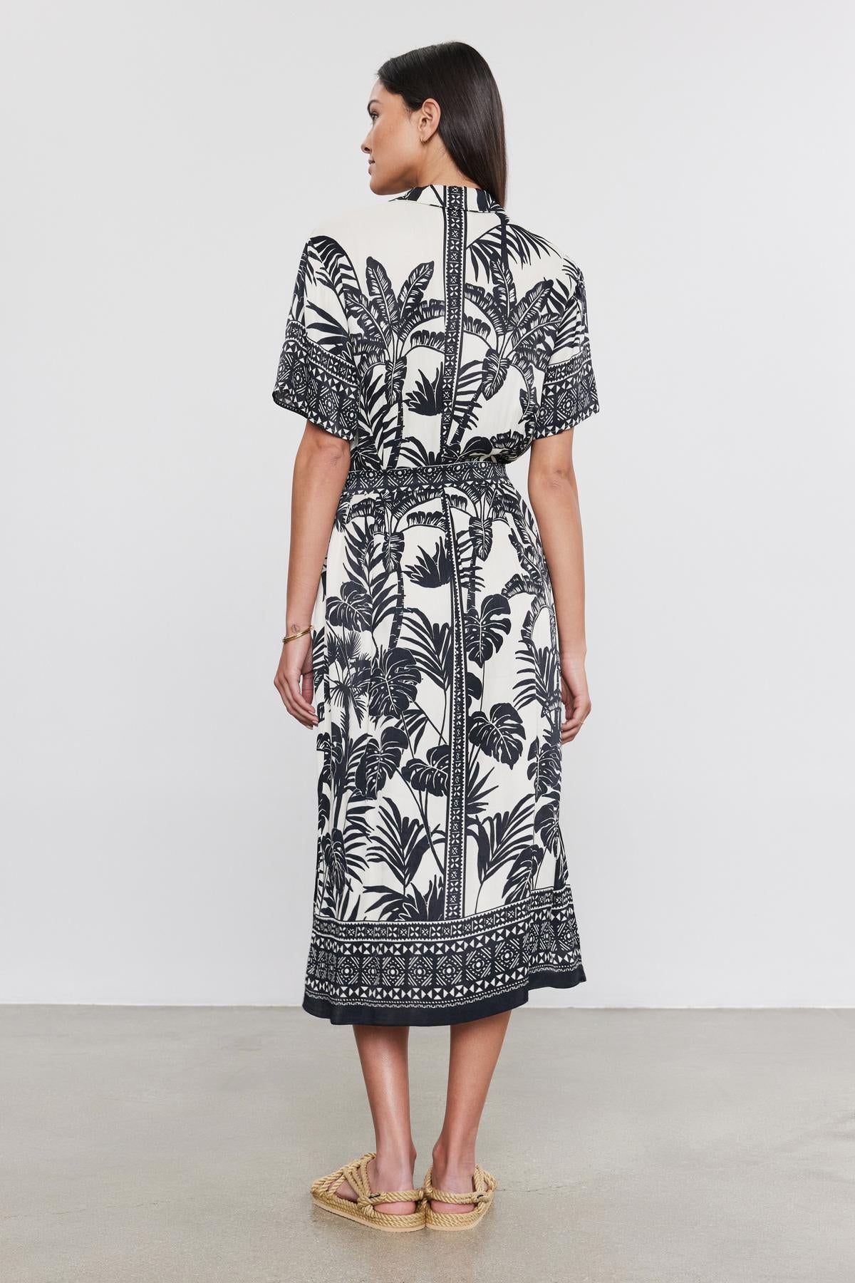   Woman in a black and white palm print FREYA DRESS by Velvet by Graham & Spencer, with a belted waist, standing in profile view against a neutral background. 