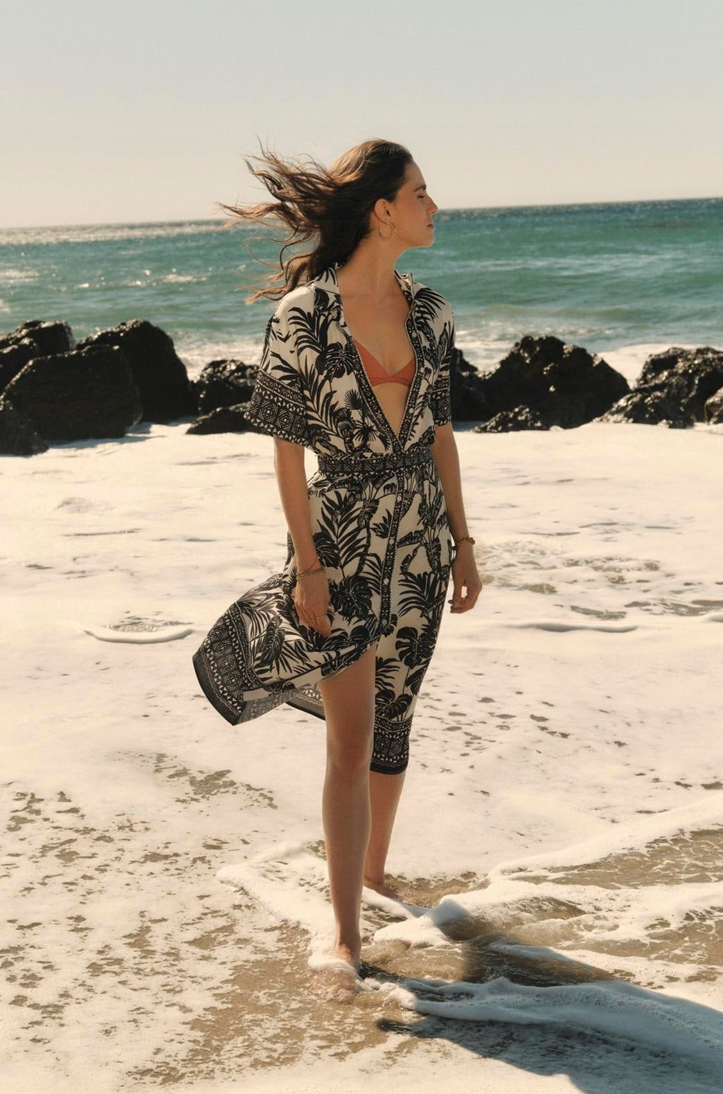 Woman in a tropical Freya dress by Velvet by Graham & Spencer standing on a sandy beach with waves touching her feet, looking out to sea, hair blowing in the wind.-36910269956289