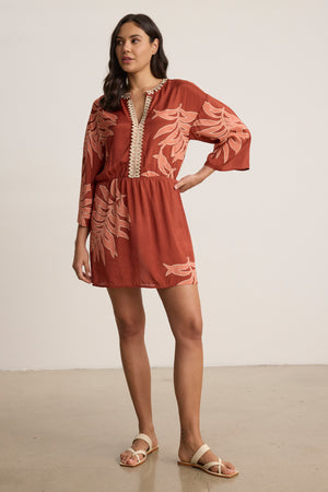A woman stands indoors wearing the PHOEBE DRESS by Velvet by Graham & Spencer—a rust long-sleeve tunic with palmetto print, v-neck, and beige sandals.
