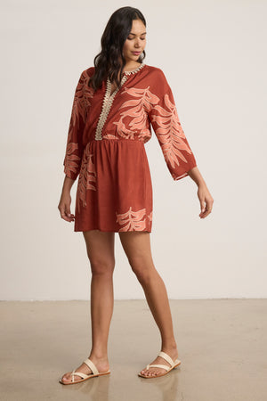 A woman stands indoors wearing the PHOEBE DRESS by Velvet by Graham & Spencer—a rust long-sleeve mini dress with an elastic waist and leafy palmetto print—paired with beige sandals, looking down with one arm extended.