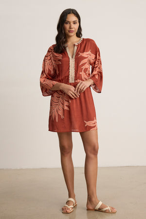A woman stands indoors on a light floor wearing the PHOEBE DRESS by Velvet by Graham & Spencer—a rust-colored, knee-length dress with beige palmetto print—paired with white sandals.