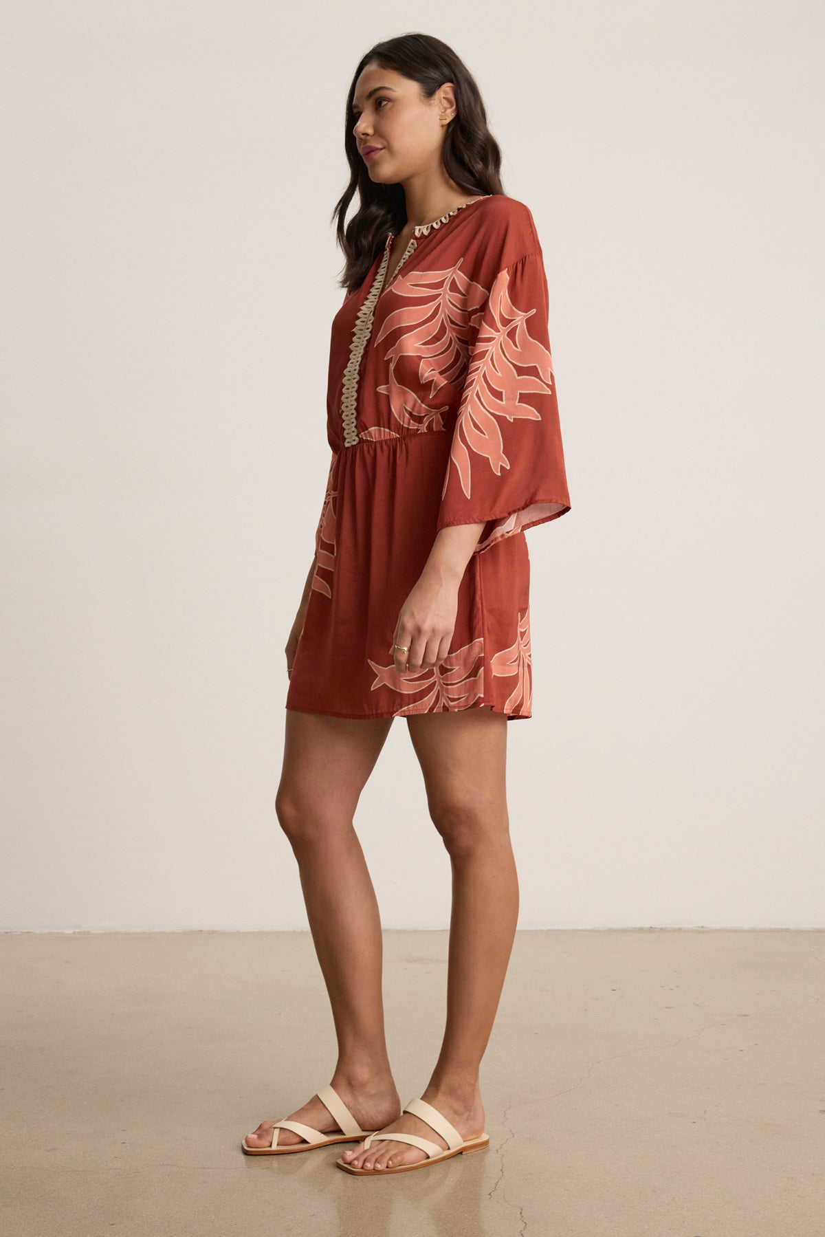 A woman stands indoors facing left against a plain background, wearing the PHOEBE DRESS by Velvet by Graham & Spencer—a rust palmetto print mini dress with long sleeves and adjustable closures—paired with light-colored sandals.-39333533581505