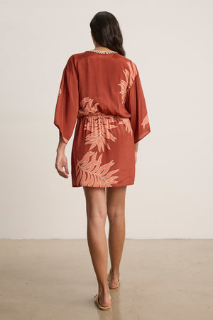 A woman faces away in the PHOEBE DRESS by Velvet by Graham & Spencer—a rust mini dress with light palmetto print—paired with sandals, set against a plain light background.
