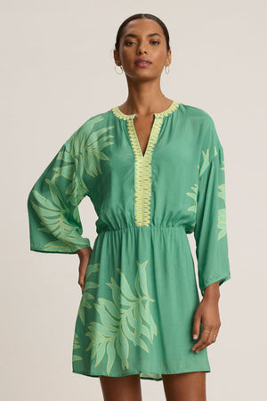 A woman stands against a plain background wearing the PHOEBE DRESS by Velvet by Graham & Spencer—a green, long-sleeved mini dress featuring a Palmetto print and gathered waist.
