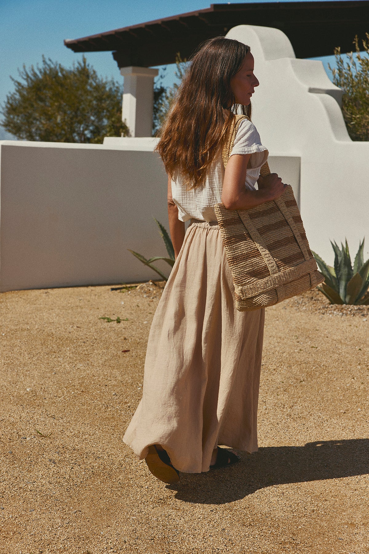 Woman in a Velvet by Graham & Spencer BAILEY LINEN MAXI SKIRT and white blouse carrying a straw bag, walking past a white stucco wall under a sunny sky.-36571158118593