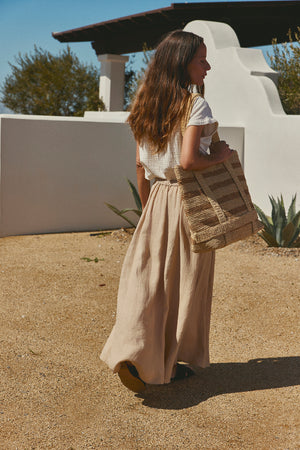 Woman in a Velvet by Graham & Spencer BAILEY LINEN MAXI SKIRT and white blouse carrying a straw bag, walking past a white stucco wall under a sunny sky.