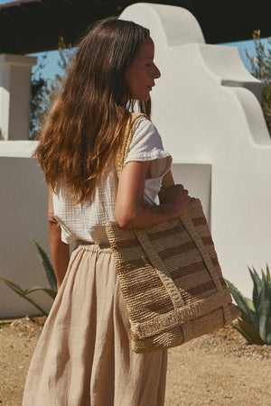 Woman standing outdoors holding a large Velvet by Graham & Spencer Raffia Straw Traveler Bag, wearing a neutral-toned blouse and skirt, with her long hair flowing, near a white stucco building.