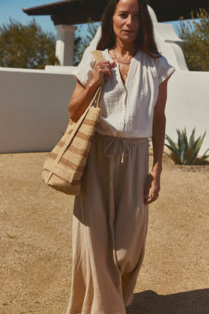 A woman in a Pamela Cotton Gauze Button-Up Top by Velvet by Graham & Spencer and beige pants carrying a straw tote bag stands in a sunny courtyard with a white wall and cactus in the background.