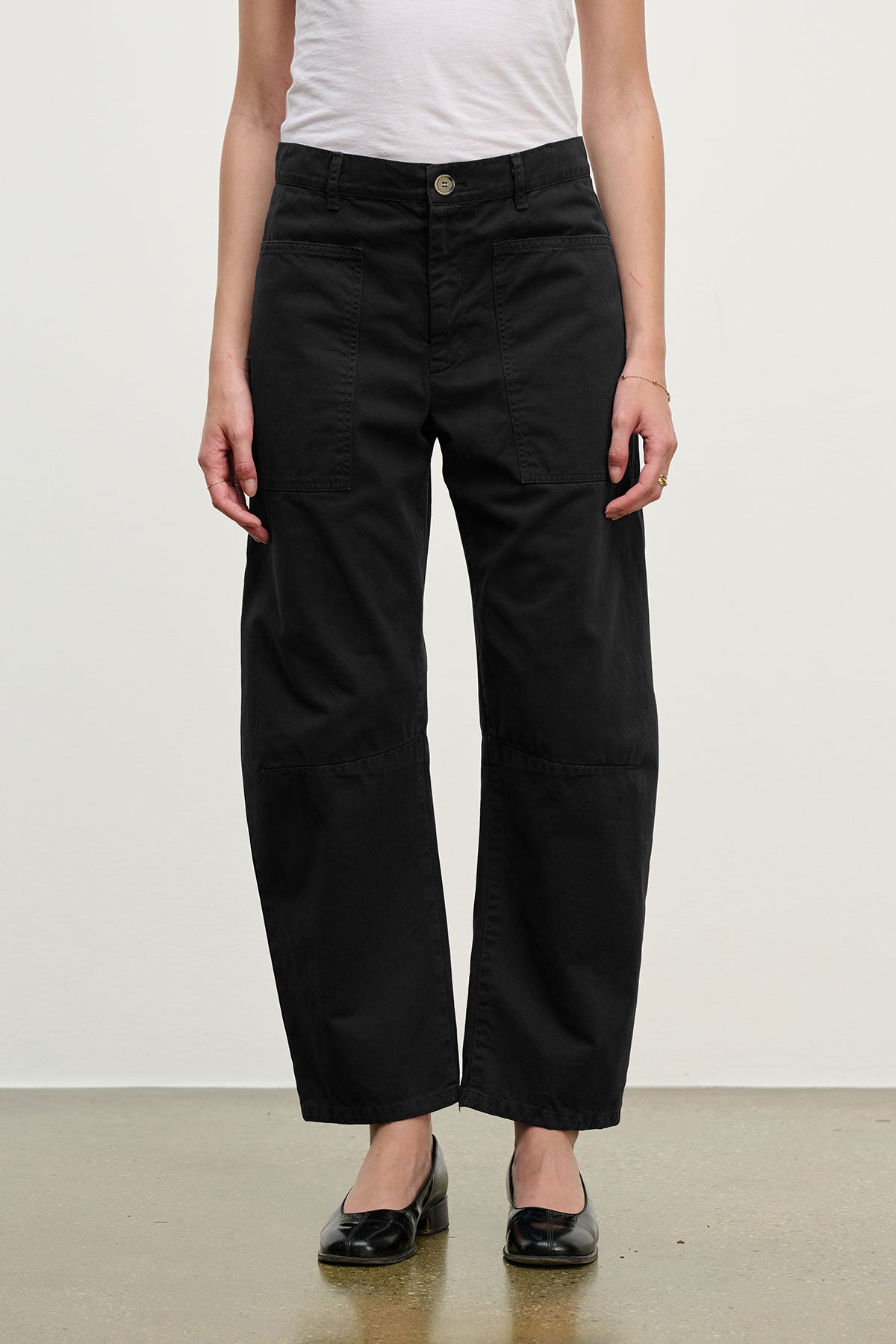 A person wears a white t-shirt, Velvet by Graham & Spencer's BRYLIE SANDED TWILL UTILITY PANT in black with patch pockets, and black flat shoes, standing against a plain background.