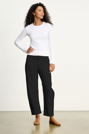 A person stands indoors in a long-sleeve white shirt and Velvet by Graham & Spencer's BRYLIE SANDED TWILL UTILITY PANT—black cotton twill casual trousers with utilitarian patch pockets—plus tan flats, posing with one hand on their hip.