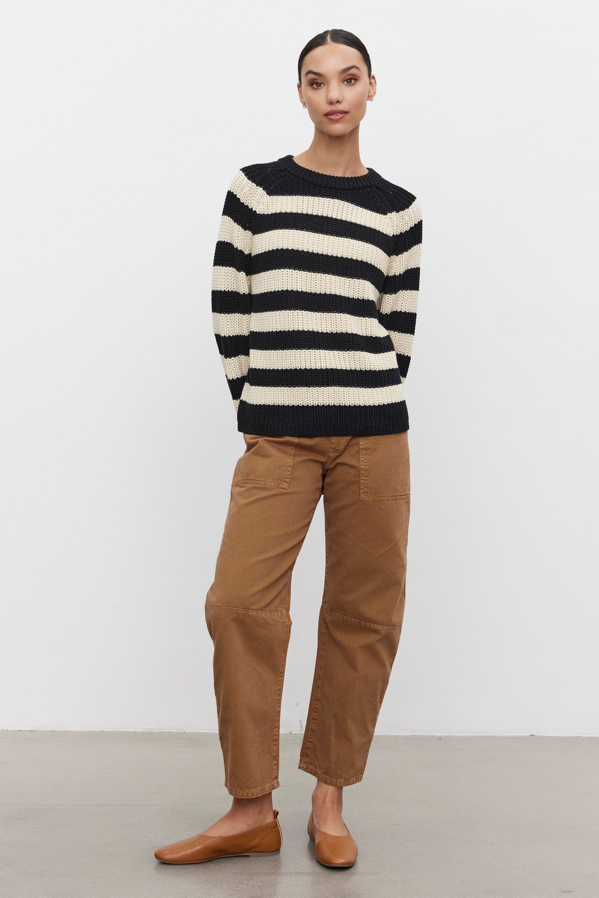   Person standing against a plain background, wearing the Velvet by Graham & Spencer OLLIE SWEATER in black and white stripes, relaxed fit brown pants, and brown flats. 