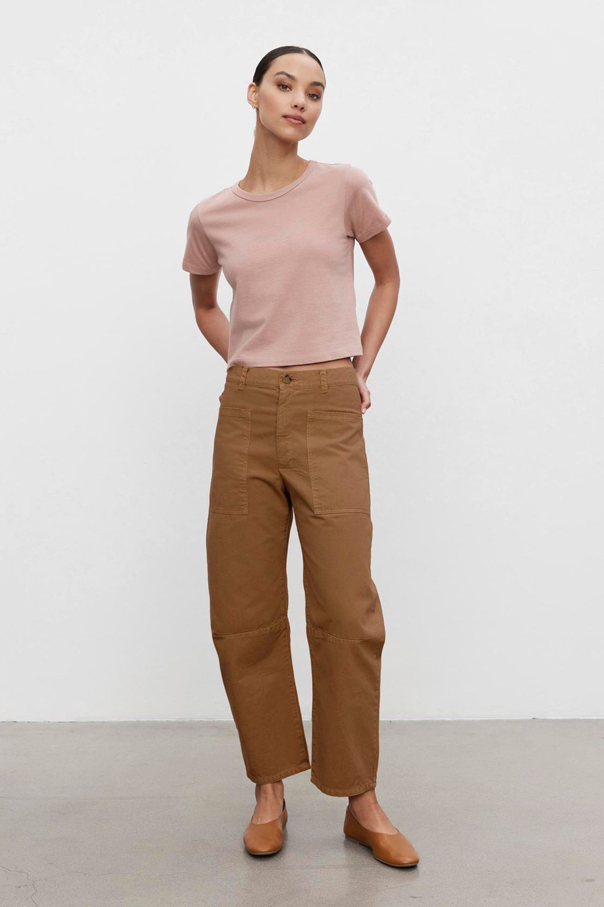   A woman wearing a light pink T-shirt, BRYLIE SANDED TWILL UTILITY PANT from Velvet by Graham & Spencer, and brown shoes stands against a plain white background with her hands in her pockets. 