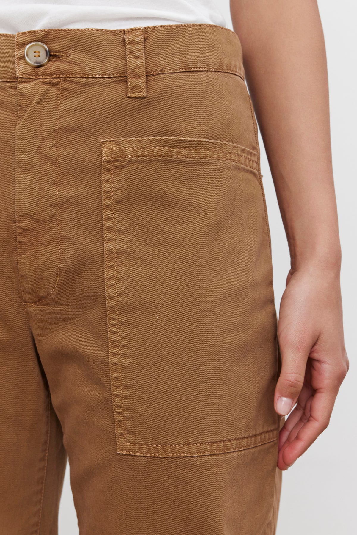 Close-up of a person wearing the BRYLIE SANDED TWILL UTILITY PANT by Velvet by Graham & Spencer, featuring a buttoned waistband and large front pocket. The casual brown cotton trouser showcases a utilitarian-inspired design. The person's hand is resting by their side, with the background plain and out of focus.-37676285591745