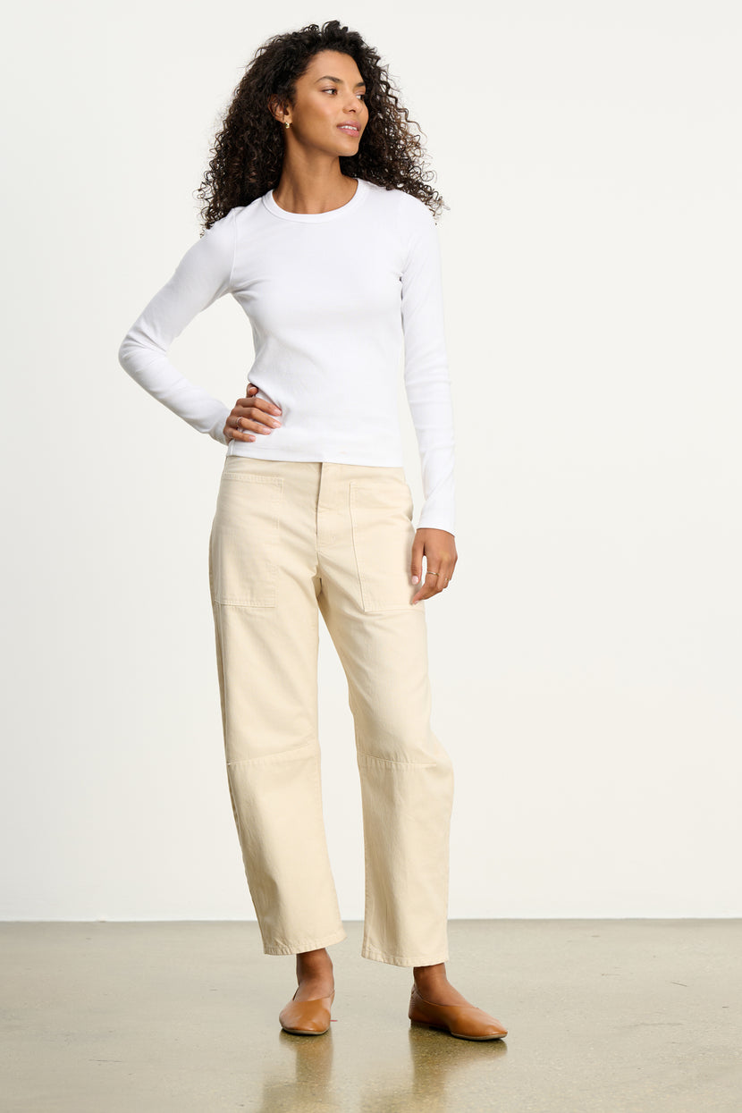 A woman stands indoors wearing a fitted white long-sleeve top and Velvet by Graham & Spencer’s BRYLIE SANDED TWILL UTILITY PANT, featuring seam details and patch pockets. She pairs the beige trousers with tan flats, her curly hair framing a softly curved silhouette.