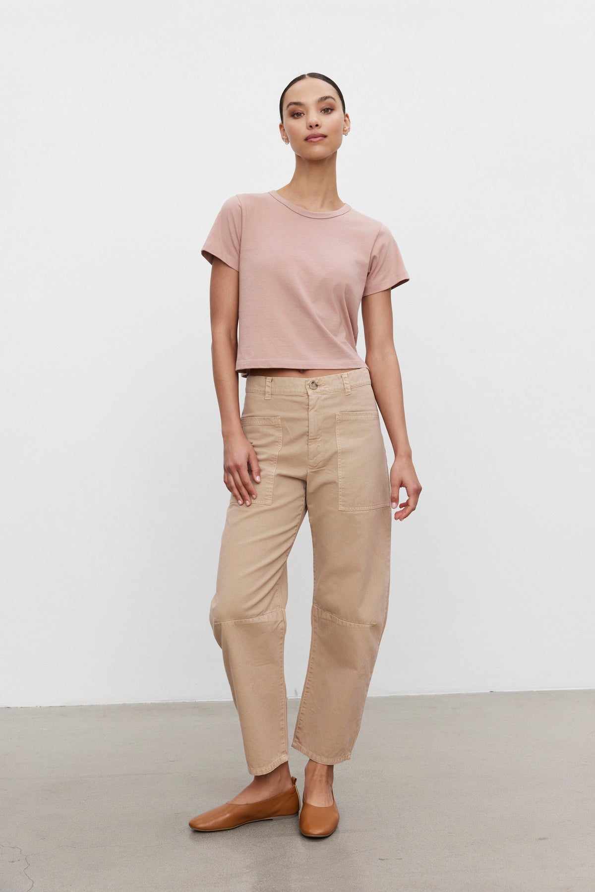   Person standing against a plain white background, wearing a light pink t-shirt paired with the BRYLIE SANDED TWILL UTILITY PANT in beige cotton twill by Velvet by Graham & Spencer, and brown shoes. 