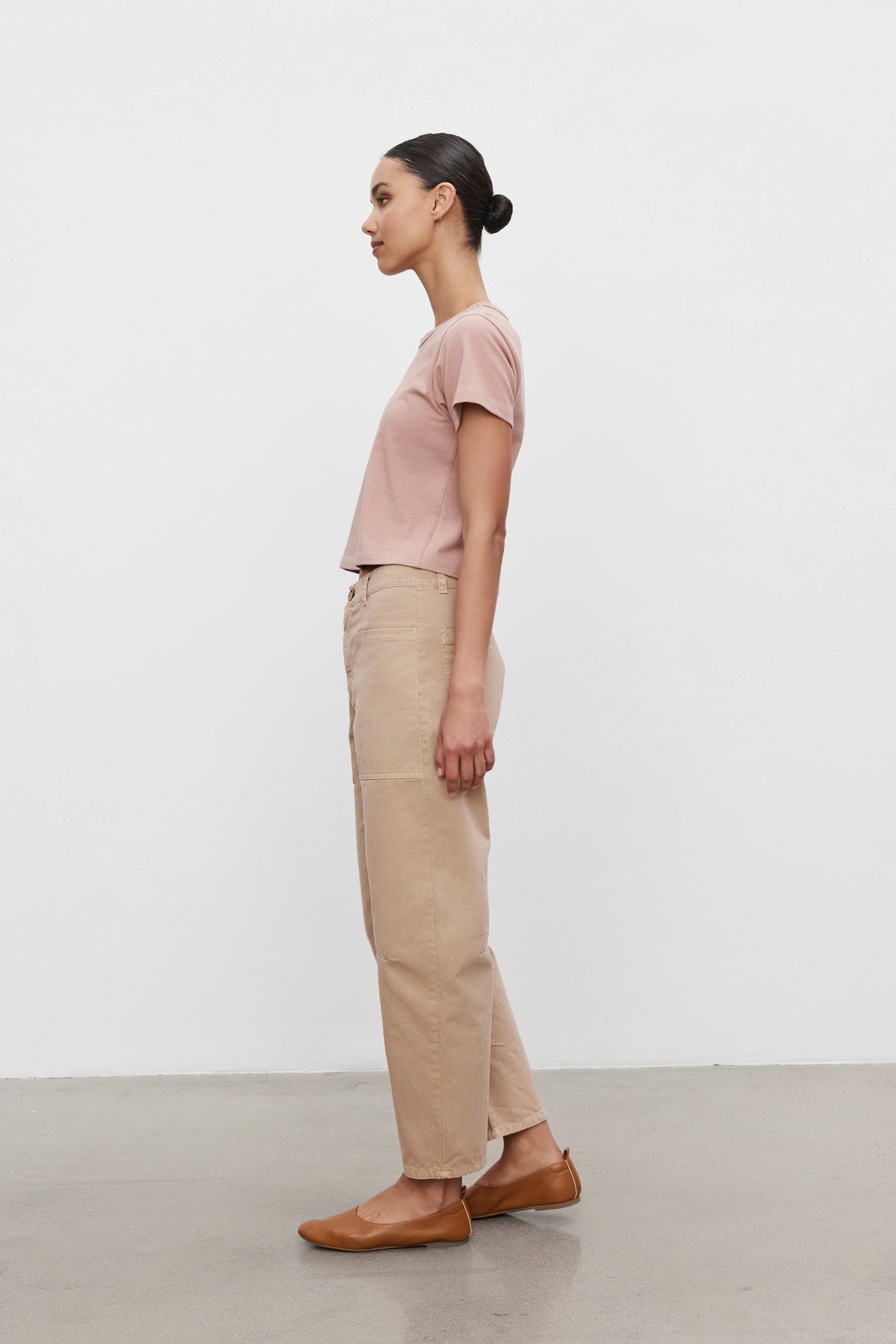 A person with tied-back hair, wearing a pink t-shirt, BRYLIE SANDED TWILL UTILITY PANT by Velvet by Graham & Spencer, and brown flats, standing in profile against a plain white background.-37629507436737