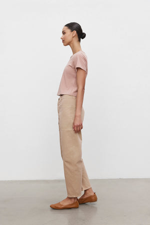 A person with tied-back hair, wearing a pink t-shirt, BRYLIE SANDED TWILL UTILITY PANT by Velvet by Graham & Spencer, and brown flats, standing in profile against a plain white background.