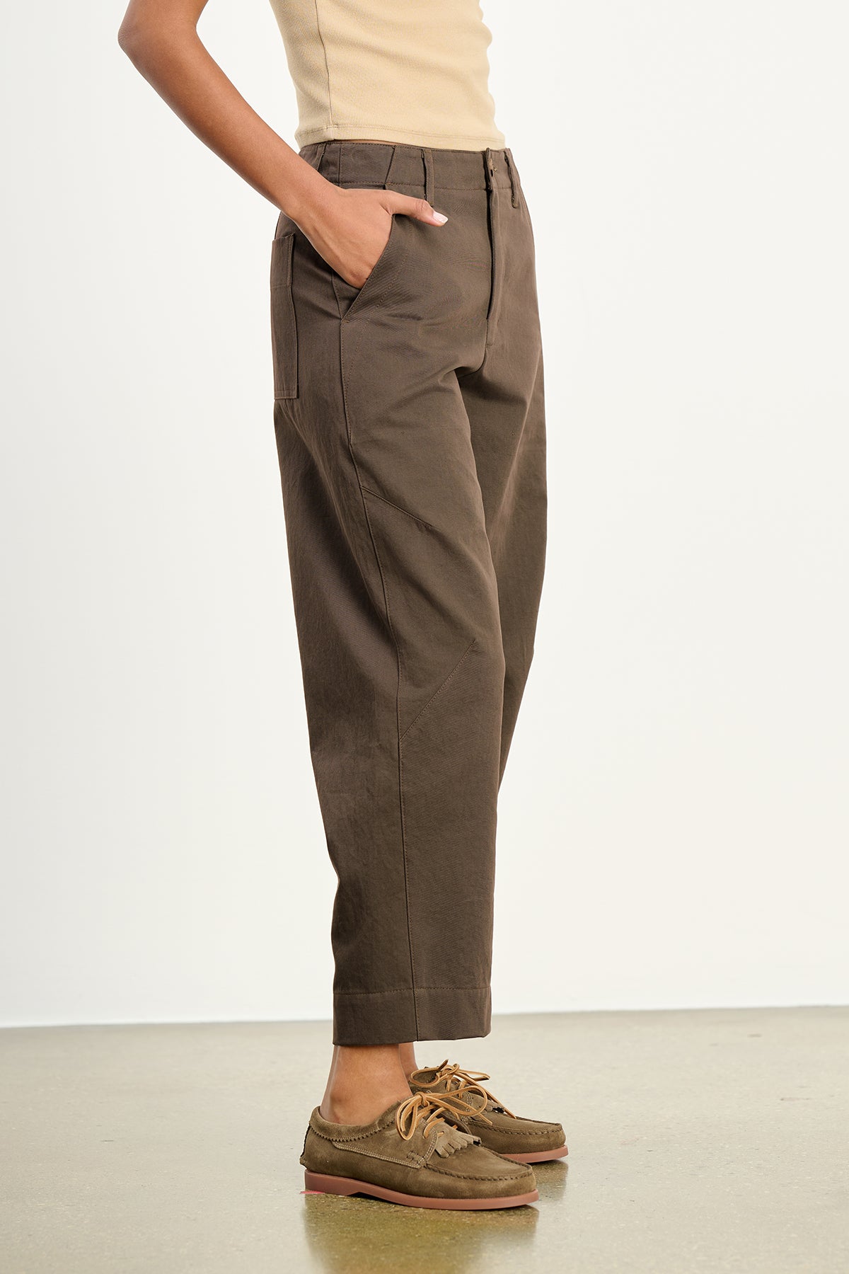 A person stands sideways with hands in pockets, wearing the DARIA PANT from Velvet by Graham & Spencer, paired with a beige top and brown boat shoes, on a light background to highlight a modern silhouette.-39559466057921