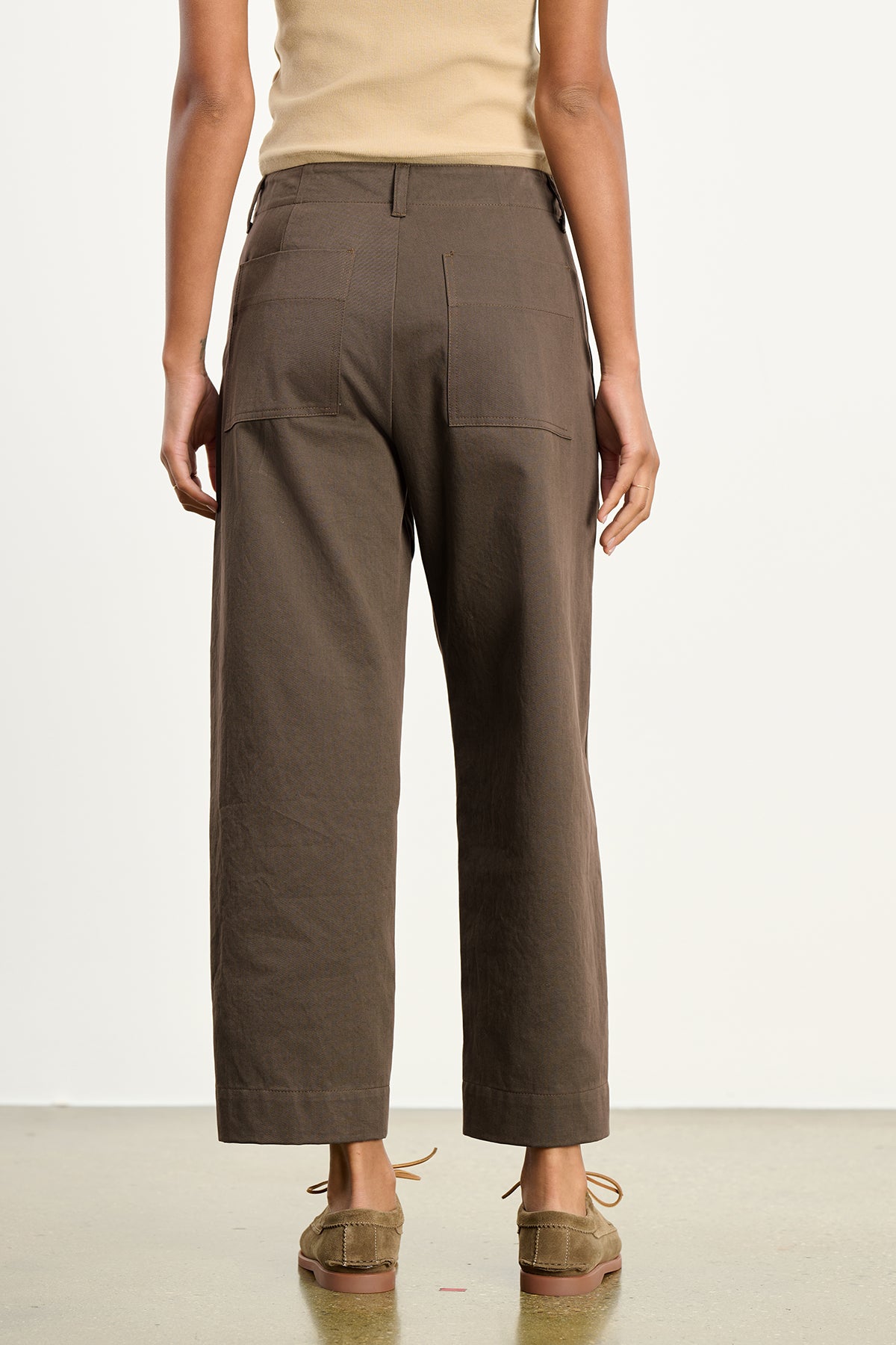   A person stands with their back to the camera, wearing Velvet by Graham & Spencer’s DARIA PANT in brown, paired with a beige sleeveless top and brown shoes against a plain background. 