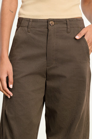 Model wears the DARIA PANT by Velvet by Graham & Spencer—brown, high-waisted, wide-leg tailored pants—styled with a beige top and hand in pocket for a sleek, modern look.