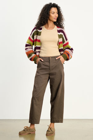 A woman stands with hands in pockets, wearing the DARIA PANT by Velvet by Graham & Spencer, a tailored ankle pant styled with a striped cardigan, beige tank top, and brown shoes against a plain background.