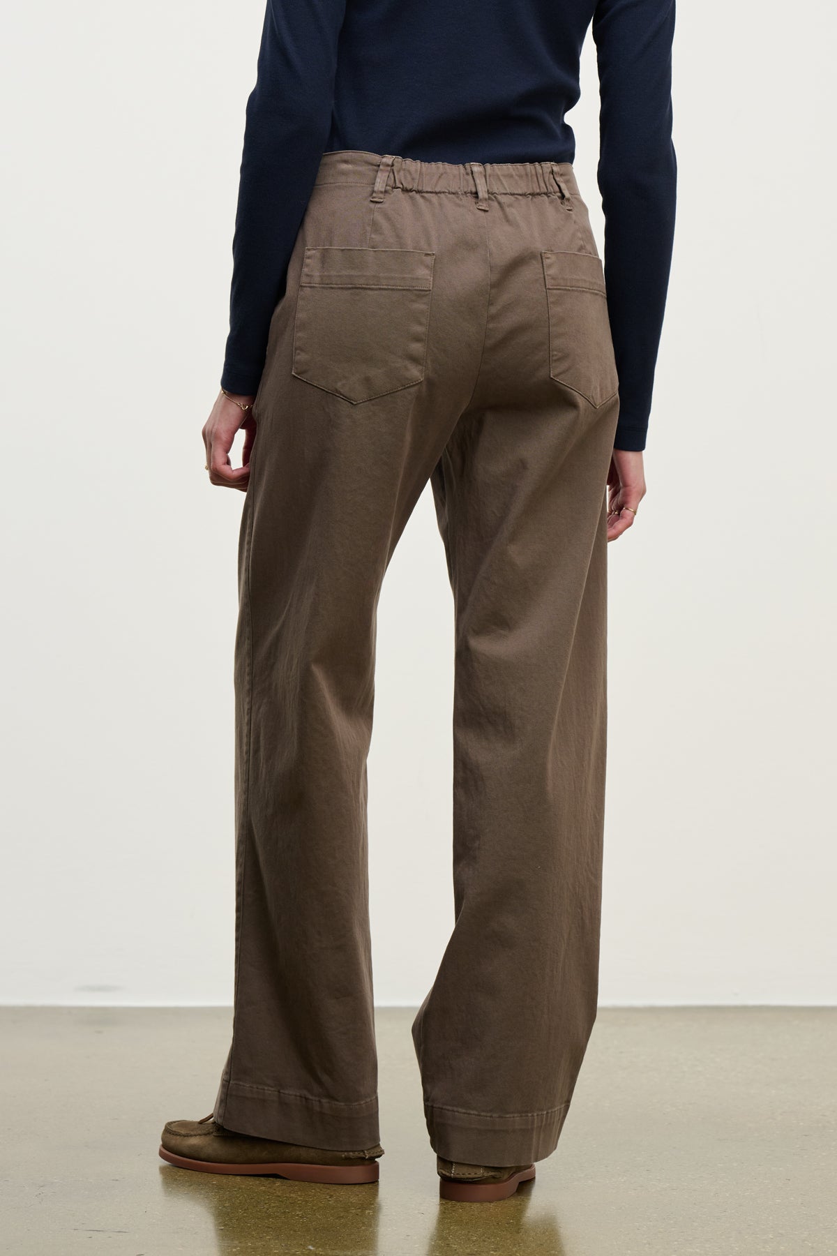   A person is shown from the back wearing the DEN PANT by Velvet by Graham & Spencer—wide-leg brown cotton twill pants with an elastic waistband—paired with a long-sleeve dark top, standing on a light floor against a plain background. 