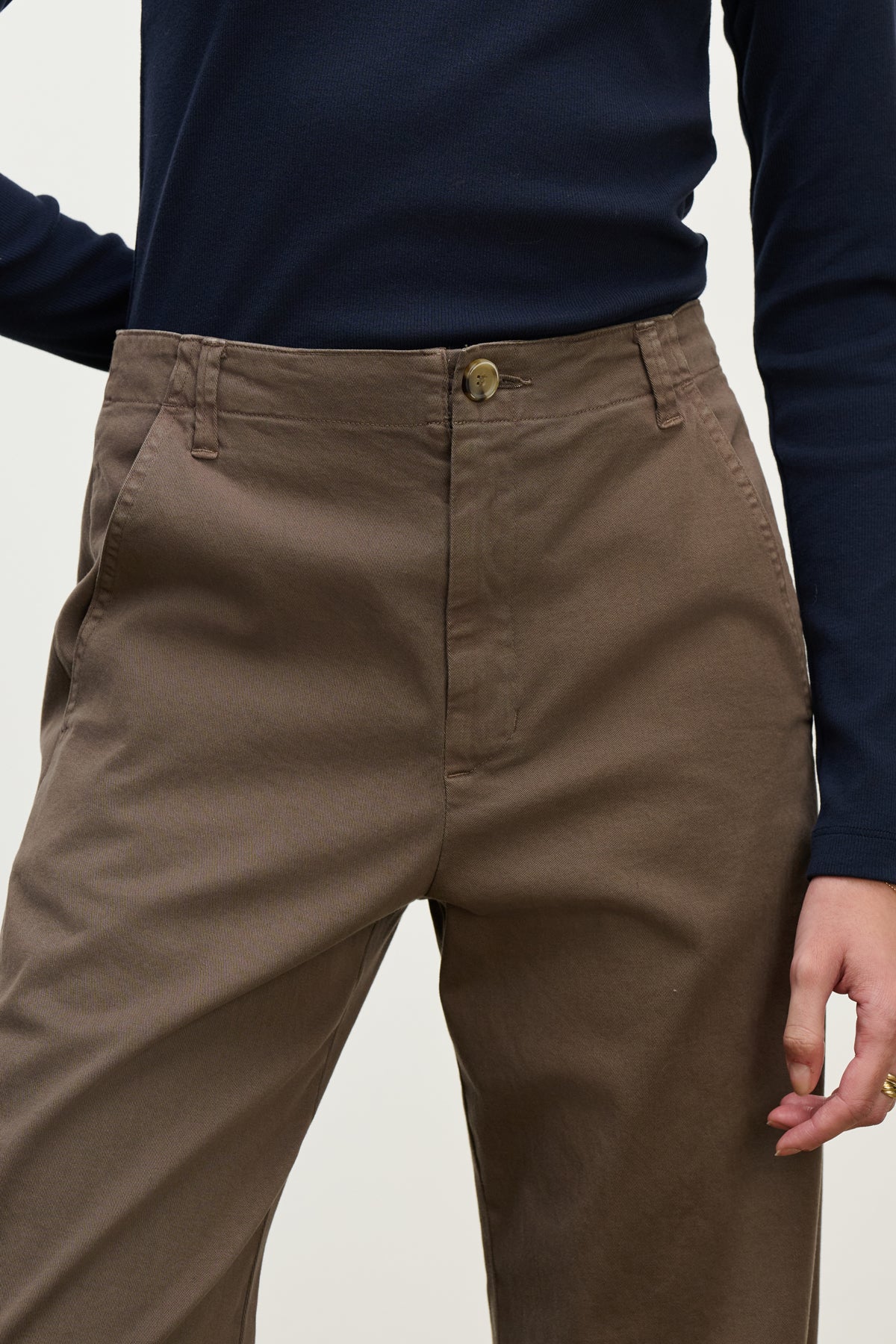   Model wears the DEN PANT by Velvet by Graham & Spencer in brown cotton twill, styled with a navy long-sleeve shirt. Shown from mid-torso to upper thighs, one hand is relaxed at their side. 
