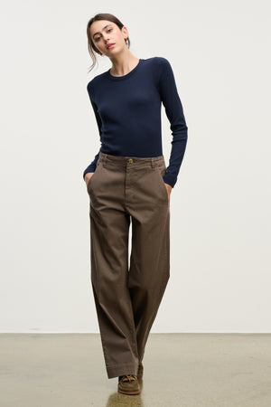 A person stands against a plain background wearing Velvet by Graham & Spencer’s DEN PANT in brown cotton twill with a wide leg and elastic waistband, paired with a navy long-sleeve shirt and brown shoes, hands in pockets.