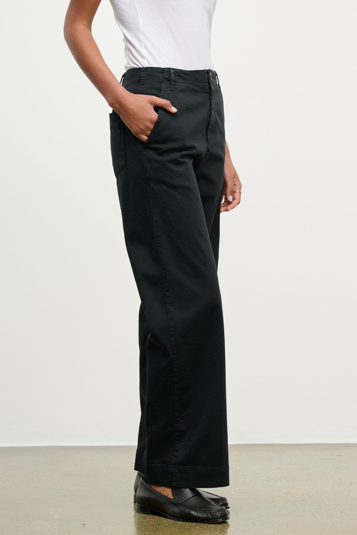 A person wearing a white top and Velvet by Graham & Spencer DEN PANT—black wide leg cotton twill with an elastic waistband—stands hands in pockets on a light-colored floor.