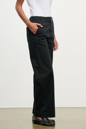 A person wearing a white top and Velvet by Graham & Spencer DEN PANT—black wide leg cotton twill with an elastic waistband—stands hands in pockets on a light-colored floor.