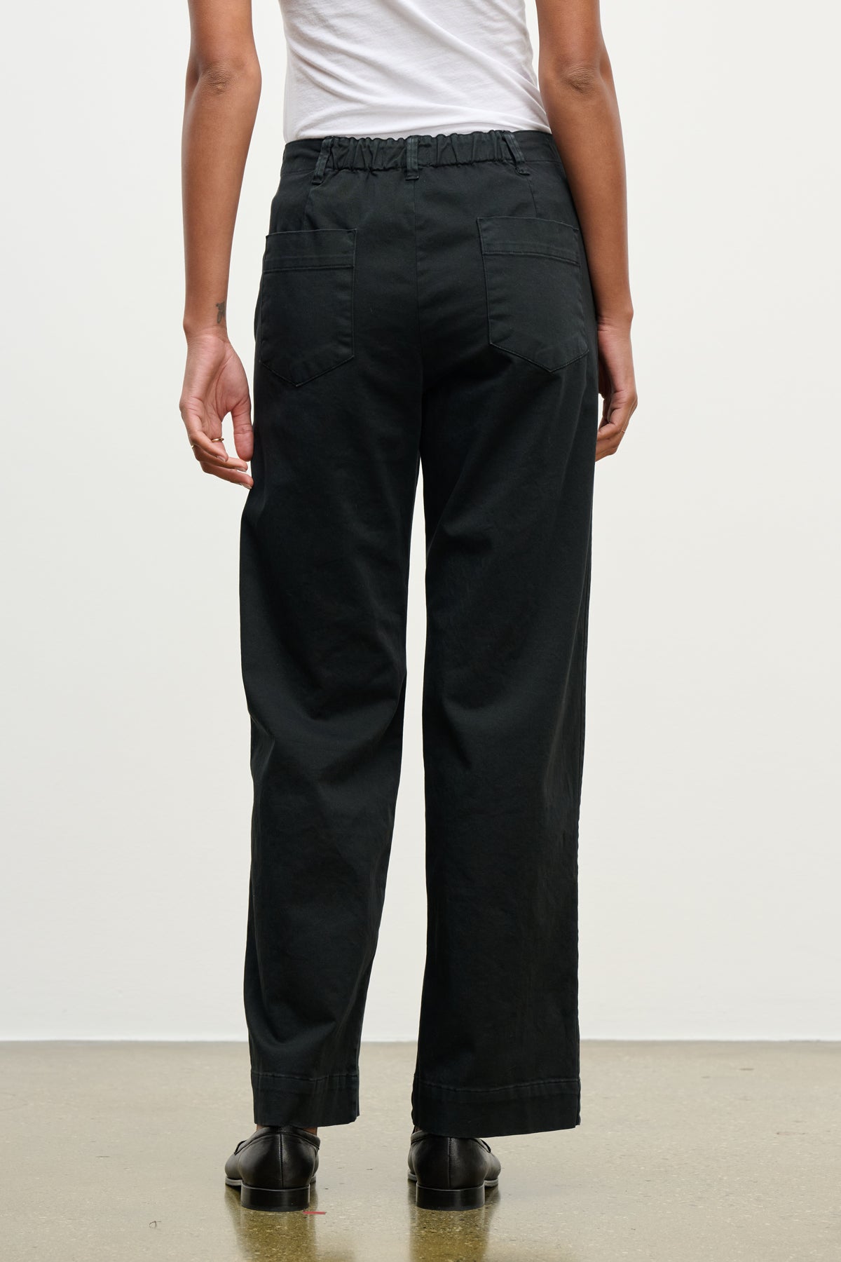 A person faces away, wearing Velvet by Graham & Spencer's DEN PANT—loose black cotton twill with an elastic waistband and back pockets—paired with a white shirt and black shoes, against a plain background.