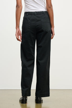 A person faces away, wearing Velvet by Graham & Spencer's DEN PANT—loose black cotton twill with an elastic waistband and back pockets—paired with a white shirt and black shoes, against a plain background.