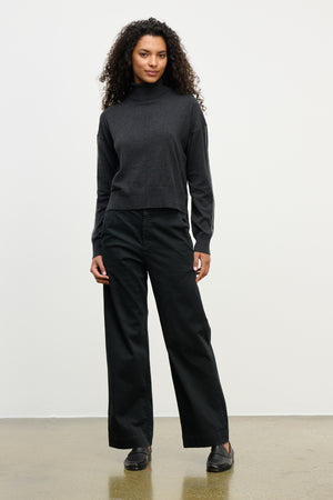 A woman stands against a plain background wearing a dark turtleneck sweater, black shoes, and the DEN PANT by Velvet by Graham & Spencer—wide leg cotton twill pants with an elastic waistband.