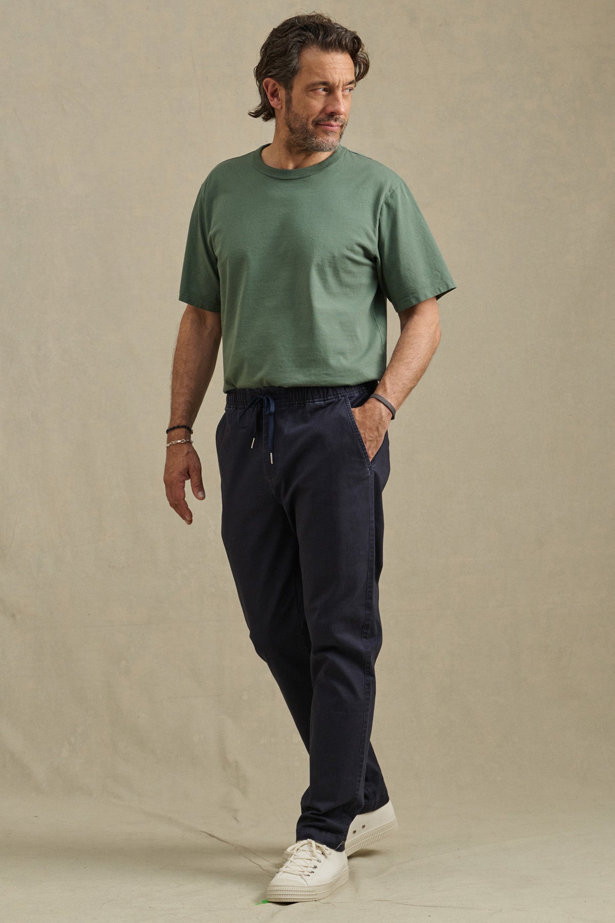 A man stands slightly turned, wearing a green tee, Velvet by Graham & Spencer's JESSE PANT in dark blue, and white sneakers against a plain beige background.-41009007689921